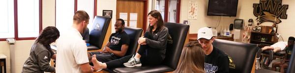 在运动训练室里，威尔逊教授坐在那里，一名学生正在包扎她的脚踝.